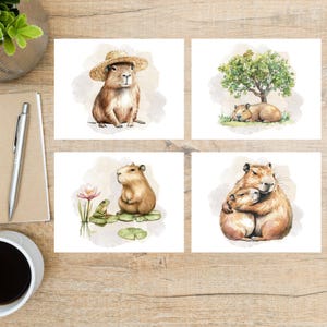 May include: Set of four watercolor art prints featuring capybaras. One print shows a capybara wearing a straw hat. Another depicts two capybaras sleeping under a tree. The other two prints show a capybara with a frog and a capybara hugging another.
