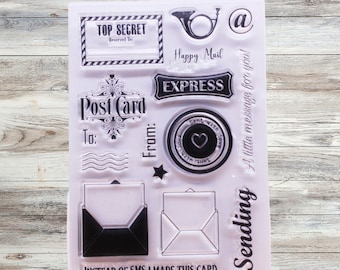 May include: A clear sheet of transparent stamps with various designs, including "Top Secret", "Post Card", "Express", "Happy Mail", "Sent with Love", and "Sending a little message for you!" The stamps are on a wooden background.
