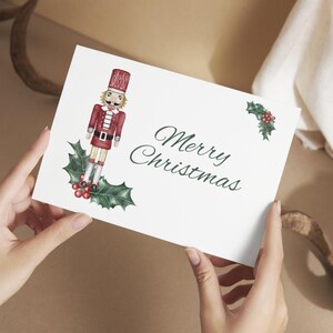 May include: A white Christmas card with the words "Merry Christmas" in green script. The card features a watercolor illustration of a nutcracker soldier in a red uniform, standing on holly with red berries. The card is held by hands.