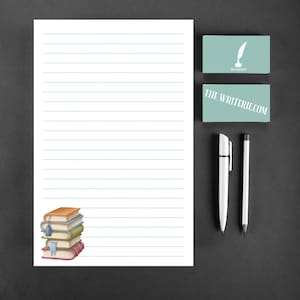 May include: A white sheet of lined paper with a stack of colorful books in the lower left corner. The paper has a blue line running down the left side. The paper is on a gray surface. There are two white pens and a business card with the text "THE WRITERIE.COM" on it.