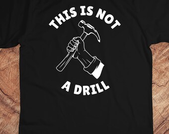 Op de afbeelding: Zwart t-shirt met een witte afbeelding van een hand die een hamer vasthoudt. De tekst "THIS IS NOT A DRILL" staat in witte, vette letters boven en onder de afbeelding.