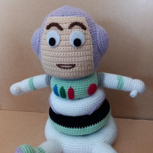 Puede incluir: Un juguete de Buzz Lightyear de la franquicia Toy Story hecho a crochet. El juguete está sentado con las piernas extendidas y tiene una cabeza morada, un cuerpo blanco y detalles verdes y negros.