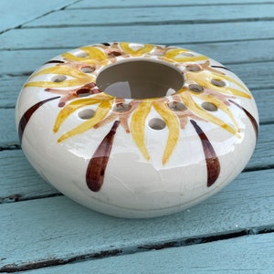 May include: A white ceramic flower frog with a yellow and brown sunflower design. The frog has multiple holes for holding flower stems.