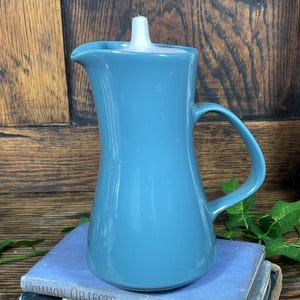 May include: A teal-colored ceramic pitcher with a handle and a white lid. The pitcher is sitting on top of two vintage blue books. The books have the titles "Common Objects of the Sea-Shore" and "Ships".