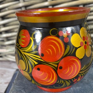 May include: A small, round, black wooden cup with a red and gold rim. The cup is decorated with a traditional Russian folk art design featuring red berries, yellow flowers, and green leaves on a black background.