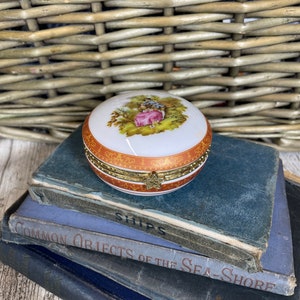 Könnte beinhalten: Eine weiße Porzellan-Schmuckdose mit goldener Verzierung und einem bemalten Bild eines Paares in einem Garten. Die Dose steht auf einem Stapel alter Bücher mit den Titeln "Ships" und "Common Objects of the Sea-Shore".