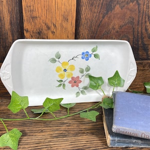 May include: A rectangular white ceramic serving dish with a floral design in yellow, pink, and blue. The dish has handles on each end and is decorated with green leaves. The background is a wooden surface.