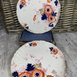 May include: Two vintage ceramic plates with a white background and a floral design. The flowers are orange, pink, and blue with green leaves. The plates have a gold rim.