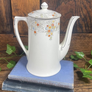 May include: A white ceramic coffee pot with a lid, decorated with floral patterns in orange, yellow, and blue. The pot has a curved handle and a spout, and is placed on top of two blue books. The pot is a vintage item.