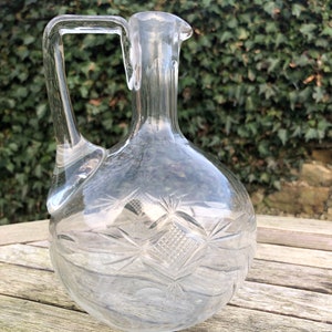 May include: A clear glass decanter with a diamond pattern etched into the surface. The decanter has a round body and a short neck with a pour spout. It has a single handle.