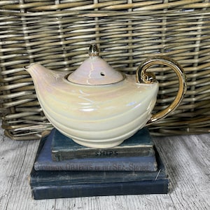 Puede incluir: Una tetera de cerámica blanca con asa y tapa doradas. La tetera tiene un acabado nacarado y está sentada sobre una pila de tres libros azules. Los libros tienen los títulos "Ships", "Common Objects of the Sea-Shore" y otro título que no es visible.