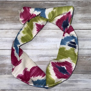 May include: A white headband with a colorful ikat pattern in shades of pink, green, blue, and navy. The headband is tied in a knot at the top.