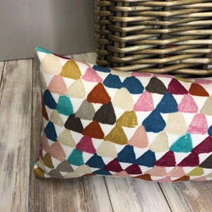 May include: A colourful throw pillow with a geometric pattern of triangles in shades of pink, blue, green, brown, and yellow. The pillow is on a light wood floor.