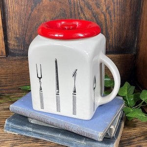 Puede incluir: Un recipiente de cerámica blanco para utensilios de cocina con tapa roja. El recipiente es cuadrado con asa y presenta ilustraciones negras de varios utensilios. Descansa sobre una pila de libros antiguos con cubiertas azules.