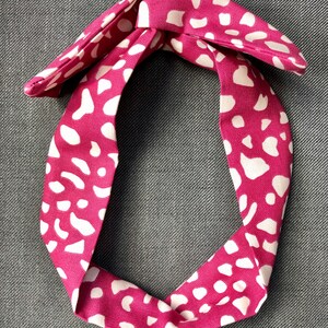 Op de afbeelding: Een roze en wit gestreept hoofdband met een geknoopte strik. De hoofdband is gemaakt van stof en heeft een witte achtergrond met roze stippen.