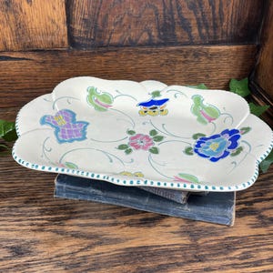 Puede incluir: Una fuente decorativa de cerámica blanquecina con borde festoneado. La fuente está adornada con coloridos diseños florales y abstractos en azul, rosa, verde y morado. Una fina línea verde punteada adorna el borde. La fuente descansa sobre un libro antiguo.
