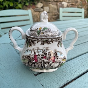 Puede incluir: Un azucarero de cerámica blanca con tapa, que representa una escena de caza con jinetes y perros en un entorno boscoso. El azucarero tiene un patrón floral marrón, verde y rojo.