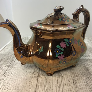 May include: A copper teapot with a floral design in shades of pink, blue, and green. The teapot has a shiny, metallic finish and a rounded body.