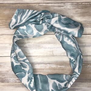 May include: A blue and white patterned headband with a knotted top. The fabric features a watercolor-style design of large leaves and foliage. The headband is shown on a light wood-grain surface.