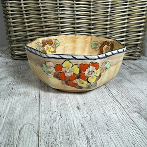 Puede incluir: Un cuenco de cerámica octogonal vintage con un diseño floral. El cuenco está decorado con flores amarillas, naranjas y rojas sobre un fondo crema. El cuenco tiene un borde negro y una franja azul.