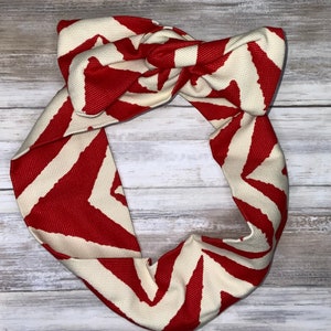 May include: A red and cream-colored headband with a knotted bow design. The headband features a bold chevron pattern in red and cream, creating a striking visual contrast. The fabric has a textured appearance.