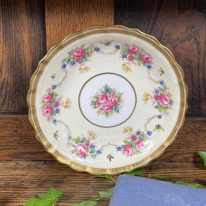 Puede incluir: Un plato vintage de color crema con un borde dorado festoneado. El plato presenta un ramo floral central y está adornado con rosas rosas, flores azules y amarillas y mariposas. Una pieza clásica para exhibir o cenar.