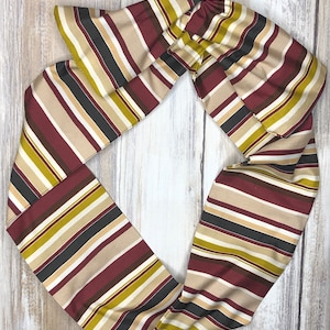 May include: A striped fabric headband in shades of burgundy, beige, black, and gold. The headband is tied in a knot at the top.