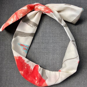 Peut inclure: Un bandeau en tissu blanc avec un imprimé floral rouge, gris et bleu. Le bandeau est noué en haut.