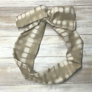 Può includere: Un cerchietto beige e bianco con un motivo a quadrati in tie-dye. Il cerchietto è annodato nella parte superiore.