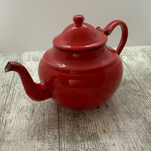 May include: A red enamel teapot with a lid and a handle. The teapot is sitting on a white wooden surface.