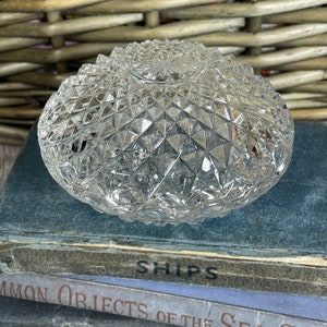 Op de afbeelding: Een heldere, diamantgeslepen glazen kom met een gestructureerd oppervlak. De kom is rond en ligt op een stapel vintage boeken. Het woord "SHIPS" is zichtbaar op de rug. De achtergrond omvat een gevlochten mand.