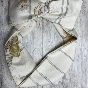 May include: A white fabric headband with a gray and gold floral pattern. The headband is tied in a knot at the top.