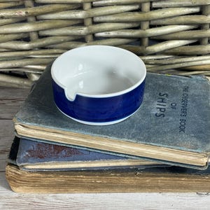 Puede incluir: Un cenicero de cerámica azul y blanco con una sola ranura para cigarrillos. El cenicero está sobre una pila de libros antiguos.