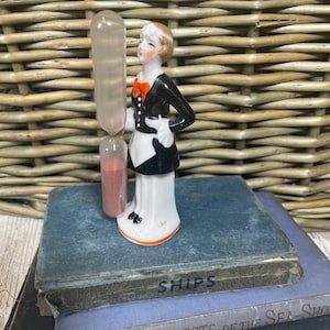 May include: A porcelain figurine of a woman in a black and white dress holding a small hourglass with red sand. The figurine is standing on a stack of books with the title "SHIPS" visible on the top book.