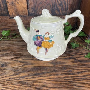 May include: Cream-colored ceramic teapot with a lid and handle. The teapot features a painted scene of two figures in period clothing dancing. The design includes embossed scrollwork and a curved spout.