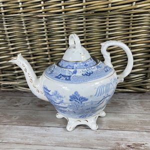 May include: A blue and white porcelain teapot with a gold trim and a Chinese landscape design. The teapot has a round body and a lid with a knob. It sits on three feet.
