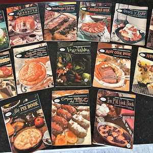 May include: A collection of vintage cookbooks with colorful covers featuring various food themes, including Christmas, desserts, casseroles, quick and easy meals, appetizers, breads and sandwiches, vegetables, meat, and pies. The cookbooks are arranged in a row on a black surface.