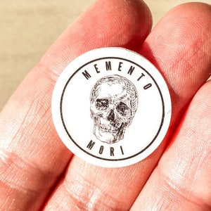 May include: A circular white sticker featuring a black skull illustration and the words "MEMENTO MORI". The sticker is held in a hand.