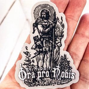 May include: Black and white sticker of a saint holding a sword and a spear. The sticker has a white background and the text "Ora pro nobis" is printed below the saint.