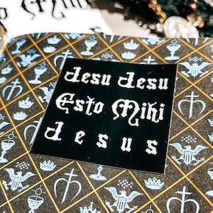 May include: A black sticker with white text that reads "Jesu Jesu Esto Miki Deus" on a background of a book cover with a repeating pattern of crowns, chalices, and crosses.