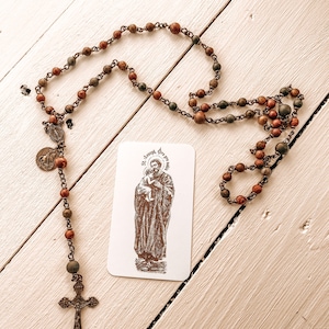May include: A wooden rosary with brown and green beads and a silver cross. The rosary is on a white wooden surface with a card that says "St. Joseph, pray for us".