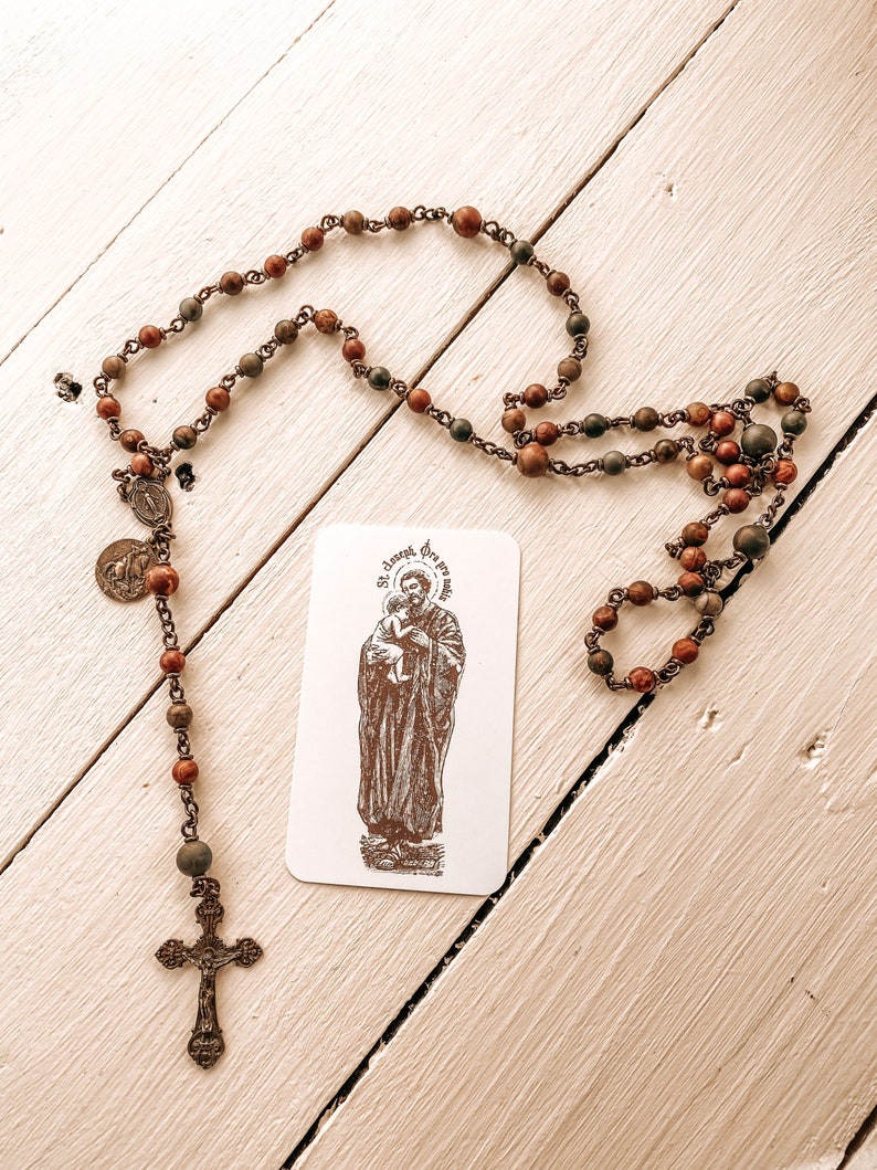May include: A wooden rosary with brown and green beads and a silver cross. The rosary is on a white wooden surface with a card that says "St. Joseph, pray for us".