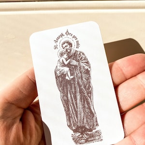 May include: A white card with a brown image of Saint Joseph holding baby Jesus. The text on the card reads "St. Joseph, Ora pro nobis".