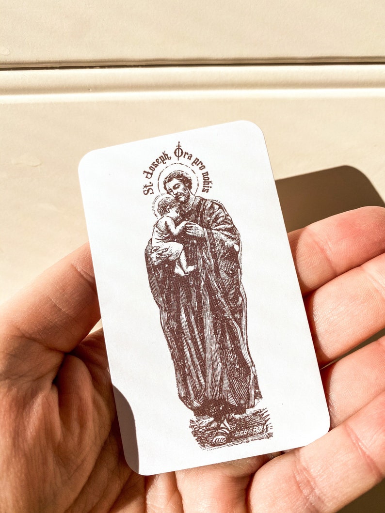 May include: A white card with a brown image of Saint Joseph holding baby Jesus. The text on the card reads "St. Joseph, Ora pro nobis".