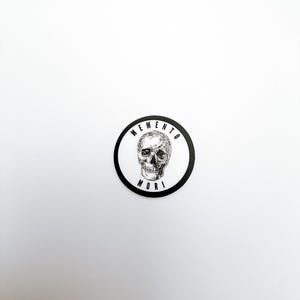 Pode incluir: Um autocolante circular com uma ilustra&ccedil;&atilde;o a preto e branco de um cr&acirc;nio humano centrado num fundo branco. O texto MEMENTO est&aacute; arqueado sobre o cr&acirc;nio e a palavra MORI est&aacute; impressa por baixo, tudo rodeado por uma borda preta espessa.