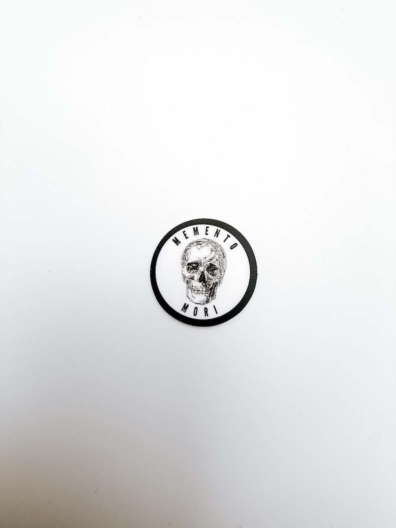 Pode incluir: Um autocolante circular com uma ilustra&ccedil;&atilde;o a preto e branco de um cr&acirc;nio humano centrado num fundo branco. O texto MEMENTO est&aacute; arqueado sobre o cr&acirc;nio e a palavra MORI est&aacute; impressa por baixo, tudo rodeado por uma borda preta espessa.