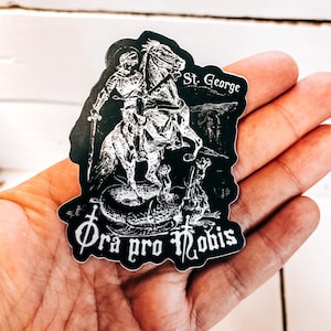 Może przedstawiać: Czarno-biała naklejka przedstawiająca rycerza na koniu zabijającego smoka. Naklejka zawiera tekst "St. George" i "Ora pro nobis" w stylu gotyckim.