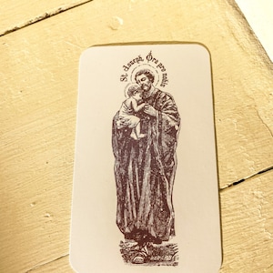 May include: A vintage-style holy card featuring a brown and white illustration of Saint Joseph holding baby Jesus. The card has the text "St. Joseph, Ora pro nobis" in a circle around the image.