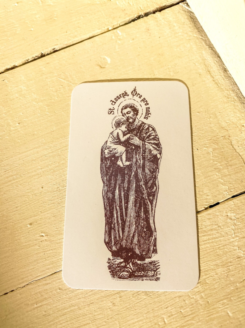 May include: A vintage-style holy card featuring a brown and white illustration of Saint Joseph holding baby Jesus. The card has the text "St. Joseph, Ora pro nobis" in a circle around the image.