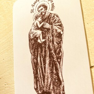 May include: A brown and white religious card with an illustration of Saint Joseph holding a baby Jesus. The card has the text "St. Joseph Ora pro nobis" in a circle around the image.